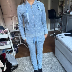 Snyggt philip plain sett i ljusblå - Snygg ljusblå hoodie och byxor med dragkedja och huva, dekorerad med breda satinband och glittriga detaljer längs ärmarna och byxben. Tröjan har ribbade muddar och fickor framtill, perfekt för en chill och sportig look.