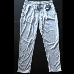 Grå mjukisbyxor Mountain Trekking L - Lätta och sköna ljusgrå mjukisbyxor från Mountain Trekking med resår och snörning i midjan. Byxorna har två sidofickor och broderad logga på ena benet. Perfekta för chill eller träning, i mjukt material som andas.