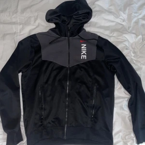 Svart och grå Nike hoodie med dragkedja - Nike hoodie i svart och mörkgrått med hel dragkedja och huva. Snyggt tryck med Nike-logga i vitt och rött på bröstet. Tröjan har långa ärmar, två fickor med dragkedja och är gjord i mjuk polyester. Perfekt för chill eller träning.