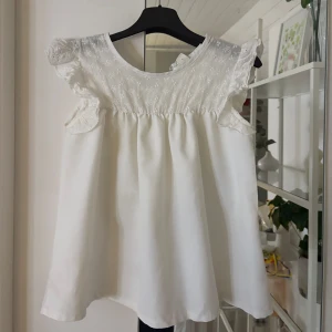 Vit blus med spets och volang - Supersöt vit blus med broderade blommor upptill och volangdetaljer vid axlarna. Blusen är ärmlös och har en knapp i nacken för enkel påklädning. Perfekt för en fräsch och romantisk look, passar till både jeans och kjol. Storlek 11 - 12 år💕