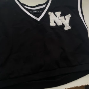 Svart croppad linnetopp med NY-detalj - Svart croppad linnetopp från PS SSTR med vit broderad NY-logga på bröstet. V-ringad med vita ränder längs halsringning och ärmöppningar. Sportig collegekänsla och perfekt för en trendig streetstyle-look.