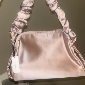 Champagnefärgad axelväska från Sensai - Superfin axelväska i champagnefärgad satin från Sensai. Väskan har en unik rynkad axelrem och stilren, mjuk form. Perfekt för dig som gillar en elegant och trendig look. Enkel att matcha med olika outfits och ger en lyxig känsla.