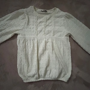 Vit strukturerad blus från C&A - Supersöt vit blus från C&A Premium i storlek 128. Blusen har långärmade puffärmar, rund hals och ett strukturerat tyg med vågig yta. Dekorativ söm framtill och resår i nederkant ger en snygg passform. Perfekt för dig som gillar stilrena och feminina plagg.