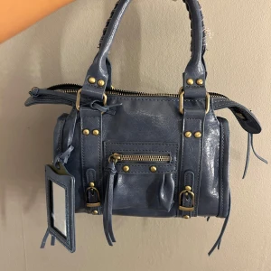 Blå handväska med detaljer - Snygg blå handväska i skinnimitation med guldfärgade metalldetaljer, dragkedjor och spännen. Väskan har dubbla handtag, en liten ficka framtill med dragkedja och en spegel i matchande fodral.