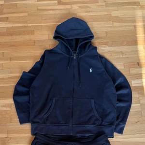 Svart hoodie från Polo Ralph Lauren - Klassisk svart hoodie från Polo Ralph Lauren med dragkedja, huva med snörning och känguruficka. Ikoniska broderade loggan på bröstet. Mjuk bomullsblandning som är perfekt för chill dagar. Passar dig som gillar stilren och enkel streetwear.