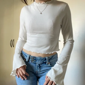 Vit  långärmad topp - Supersöt vit ribbad croppad topp med långa trumpetärmar och volangkant vid hals och ärmslut. Ribbad i tyget och croppd. Ursprungligen från NA-KD och i storlek xxs. Den har tecken på användning. 