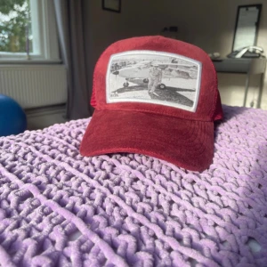 Röd keps från WON med motiv - Snygg röd keps från WON med mesh på baksidan och justerbar snapback. Framsidan har ett svartvitt motiv av ett flygplan och pilot. Kepsen har en cool vintagekänsla. Märkt 'Born in Sweden' på insidan.