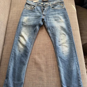 Blå Replay jeans Rocco herrmodell - Säljer ett par blå Replay jeans, modell Rocco, med snyggt slitna detaljer och urtvättning samt klassisk femficksdesign. Jeansen har normal passform, dragkedja och är tillverkade i mjukt bomullsjeansmaterial. Perfekta för dig som gillar en avslappnad men stilren look. 🙌🙌