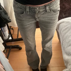 Grå bootcut jeans med detaljer - Säljer ett par grå jeans med bootcut-modell och snygga detaljerade bakfickor med flärp och knapp. Jeansen har normal passform och klassisk femficksdesign. Perfekta för dig som gillar en avslappnad men ändå trendig look.