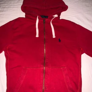 Röd hoodie från Polo Ralph Lauren - Röd hoodie från Polo Ralph Lauren med dragkedja och vit snörning i huvan. Klassisk broderad logga på bröstet och två fickor framtill. Perfekt för en avslappnad stil. Finns litet hål inne vid luvan men inget som syns vid användning.