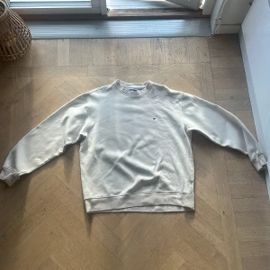Vit sweatshirt från Tommy Hilfiger - Snygg vit sweatshirt från Tommy Hilfiger med klassisk rund hals och diskret broderad logga på bröstet. Tröjan har långa ärmar och ribbade muddar vid ärmslut och nederkant. Perfekt för en clean och stilren look.