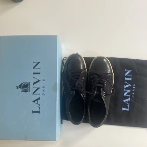 Svarta sneakers från Lanvin i mocka och skinn - Snygga svarta sneakers från Lanvin med ovandel i mocka och tå i blankt skinn. Skorna har vita platta sulor och svarta snören. Klassisk och stilren design med metallfärgade öljetter och diskreta detaljer. Kommer med originalkartong och dustbag.