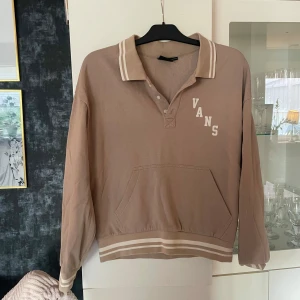 Beige pikétröja från Vans - Snygg beige pikétröja från Vans med vita ränder på krage, ärmslut och midja. Tröjan har lång ärm, klassisk krage med knappar. Retro tröja, tydligt använd men väl omhändertagen. 