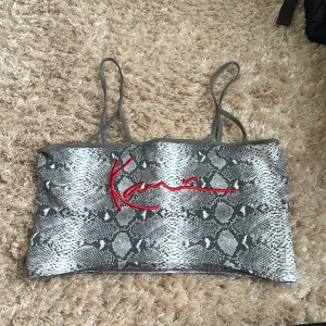 Snakeprint topp från Karl Kani - Säljer en grå och vit snakeprint croptop från Karl Kani med tunna axelband och röd broderad logga framtill. Toppens material är stretchigt och passar perfekt till dig som vill sticka ut med ett coolt mönster och streetkänsla.