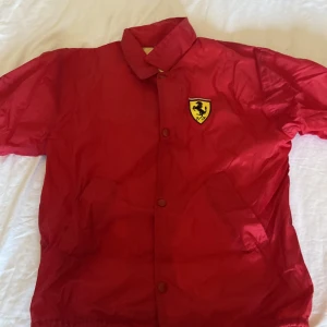 Röd Ferrari vindjacka med logga - Riktigt snygg röd vindjacka från Ferrari med klassisk gul Ferrari-logga på bröstet. Jackan har krage, tryckknappar framtill och elastisk nederkant. Perfekt för dig som vill sticka ut och visa racing-vibbar. Materialet är lätt och känns sportigt.