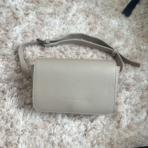Beige midjeväska från Smith & Canova - Snygg beige midjeväska från Smith & Canova i stilren design. Väskan är i slätt skinn med justerbart axelband och har ett huvudfack med dragkedja 2 små innerfack samt lila innerfoder. Perfekt storlek för det viktigaste och passar till många outfits.