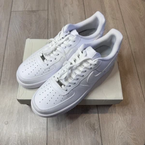 Nike Air Force 1 helvita sneakers - Klassiska Nike Air Force 1 sneakers i helvitt skinn med perforerad tå, rund tå och platt sula. Snygg och tidlös modell med snörning och diskret Nike-logga på hälen. Perfekta för dig som vill ha en clean och stilren look.                                     Storlek:46  Skick: Helt nya  📦 Skickas inom 1–3 dagar. 📩 För mer information eller frågor, kontakta mig gärna.