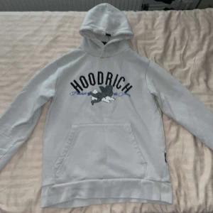 Ljusgrå HOODRICH hoodie  - Säljer en ljusgrå hoodie från HOODRICH med stor svart och blå logga framtill och coolt tryck med änglar och text på ryggen. 