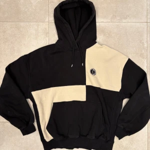 Svart och beige hoodie från District Fortysix  - Snygg hoodie från District Fortysix i svart och beige blockdesign. Tröjan har huva med snörning, stor magficka och broderad logga på bröstet. Relaxed oversize fit. (Passar S/M)