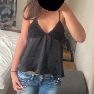 Svart satin linnetopp med spets - Supersnygg svart linnetopp i satin med tunna axelband och v-ringning. Toppen har spets och broderade blommor vid bysten samt knyt i ryggen för extra detalj. Perfekt att styla med jeans eller kjol för en trendig look.