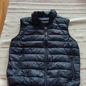  Mörkblå väst  från Polo Ralph Lauren - Säljer en mörkblå dunväst från Polo Ralph Lauren med ståkrage och dragkedja framtill. Västen har två fickor med dragkedja och en liten röd broderad logga på bröstet. Perfekt för lager-på-lager under kyliga dagar. Storlek Medium
