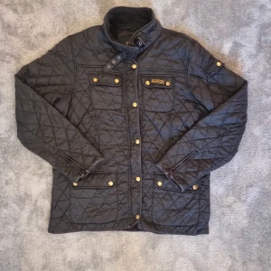 Svart quiltad jacka från Barbour - Säljer en svart quiltad jacka från Barbour med guldiga knappar och hög krage med spänne. Jackan har fyra fickor framtill och Barbour-logga på bröstet. Perfekt för kyliga dagar och snygg till många outfits                                                     en dragkedja är borta och går inte att stänga med de men man kan stänga med jnapparna. Pris kan sänkas