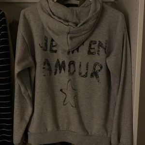 Grå hoodie med tryck  - Grå hoodie med texten 'Je m'en amour' och en stjärna tryckt både fram och bak. Mjuk bomullskänsla, huva och ribbade muddar vid ärmslut och nederkant. Liten synlig fläck på ryggen