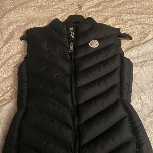 Svart dunväst från Moncler - Säljer en svart dunväst från Moncler med dragkedja framtill och hög krage. Västen har ett klassiskt Moncler-märke på bröstet och är perfekt för lager-på-lager-stil. Snygg och tidlös design som passar till mycket.