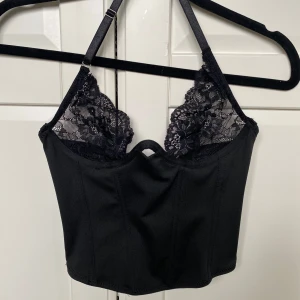 Svart spetskorsett - Säljer en svart korsett-topp med fina spetsdetaljer upptill och smala axelband. Toppen har en croppad passform och en liten öppning framtill. Perfekt för att lyfta vilken outfit som helst! Aldrig använd 