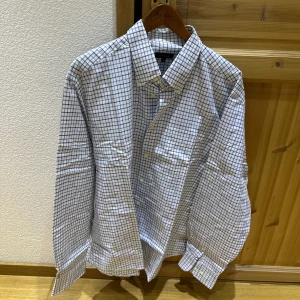 Vit rutig skjorta från Derby - Klassisk vit skjorta från Derby med blått rutmönster. Skjortan har lång ärm, knappar framtill och en bröstficka. Perfekt för dig som gillar en stilren och tidlös look.