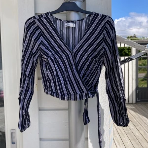Svartvit omlottopp med knyt från Hollister - Säljer en svartvit omlottopp från Hollister med smala vertikala ränder. Toppen har långa ärmar, croppad passform och knyt i midjan. Perfekt för dig som gillar en trendig och stilren look.