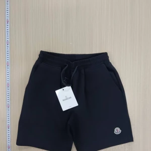 Svarta shorts från Moncler - Svarta shorts från Moncler med elastisk midja och snörning. Klassisk design med två sidofickor och Moncler-logga på vänster ben. Tillverkade i mjukt material som passar perfekt för avslappnade dagar.