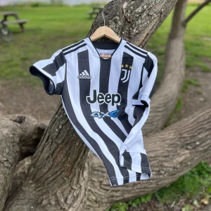 Juventus Hemmatröja 2021/2022 Pogba #10 - Juventus hemmatröja med svarta och vita ränder, Adidas-logga och Jeep som sponsor. Pogba och nummer 10 på ryggen. Klassisk V-ringning och tre stjärnor ovanför klubbmärket. Texten '10 Years At Home' i nacken.