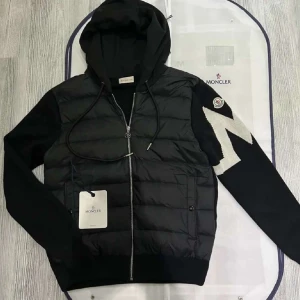 Moncler cardigan - Svart pufferjacka från Moncler med huva och dragkedja framtill. Jackan har stickade ärmar med vit zigzagdetalj och Moncler-logga på ena ärmen. Fodrad kropp och två fickor framtill. Perfekt för kyliga dagar.