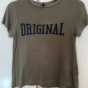 Khakigrön t-shirt med tryck från Divided - Khakigrön t-shirt från Divided med svart text 'ORIGINAL' på bröstet. Klassisk rund halsringning och korta ärmar. Perfekt för en avslappnad och cool stil.