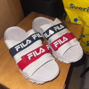 Vita sandaler från FILA med röda och marinblå remmar - Säljer ett par vita sandaler från FILA med två breda remmar i rött och marinblått, båda med stor vit FILA-logga. Sandalerna har en grov, mönstrad sula för extra grepp och är perfekta för sommaren.