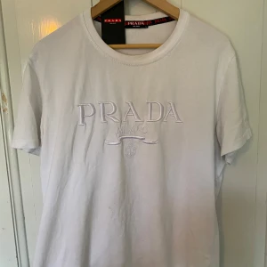 Prada tröja  - Säljer en vit t-shirt från Prada med broderad logga framtill. Klassisk rund halsringning och korta ärmar. Perfekt för dig som gillar stilrena och exklusiva plagg. T-shirten är i mjuk bomull och har en snygg passform. Står storlek L, men skulle säga den pasdar M bäst. Fick i födelsedagspresent förra året, kommer med lappar, qr koden tar dig till Prada hemsidan 💯