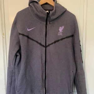 Liverpool Nike Tech Fleece - Säljer en lila tech fleece från Nike med Liverpool-logga på bröstet. Jackan har dragkedja, huva och sportig design med svarta detaljer. Perfekt för fotbollsfans som vill ha en snygg och bekväm jacka.