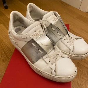 Valentino Garavani sneakers med silverdetalj - Säljer ett par vita Valentino Garavani sneakers med bred silverfärgad rem över sidan. Skorna har klassisk snörning och rund tå, samt dekorativa nitar baktill på sulan. Perfekta för dig som gillar stilrena och exklusiva sneakers. Kvitto och originalkartong finns.