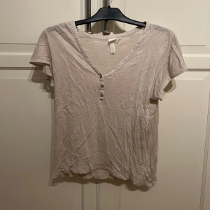 Beige linnetopp med knappar från H&M - Säljer en beige linnetopp från H&M i storlek S. Toppen har korta ärmar, v-ringning och tre dekorativa knappar framtill. Tillverkad i linne vilket gör den sval och skön. Perfekt för varmare dagar!