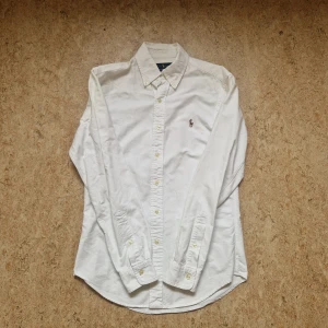 Vit skjorta från Ralph Lauren - Klassisk vit skjorta från Ralph Lauren i slim fit-modell. Skjortan har långa ärmar, knappar framtill och broderad logga på bröstet. Perfekt för en clean och stilren look. Liten fläck syns på sista bilden annars nyskick. Fläcken går troligtvis att få bort i tvätten!