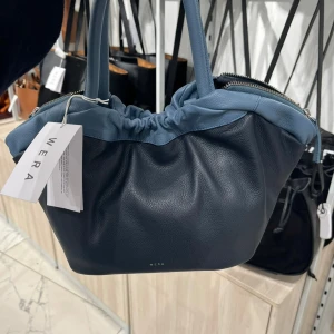 Mörkblå handväska från Wera med ljusblå detaljer - Mindre tote bag i med elegant design i två färger. har rejäla handtag i läder och stängs med magnetisk tryckknapp i mitten och dragkedjor på utsidan om handtagen. Huvudfacket har en ficka med dragkedja samt korthållare med plats för fyra kort. Löstagbart och justerbart axelband medföljer.   Mått: Längd: 40/20 cm Höjd (med handtag): 33 cm. Bredd: 16cm