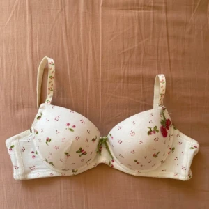 Vit bh med jordgubbsmönster - Supersöt vit bh med små jordgubbar och blommor som mönster. Axelbanden är justerbara och har samma mönster som bh:n. Perfekt för dig som gillar gulliga detaljer och vill ha något unikt i underklädeslådan.