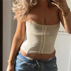 Vit korsett-topp - Säljer en snygg vit korsett-topp med lätt glansig yta och diskret mönster. Toppen är axelbandslös och har en tight passform som framhäver midjan. Perfekt att styla med jeans eller kjol för en trendig look. Maidenform! Storlek xs men skulle även passa s