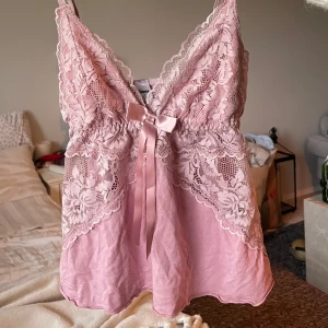 Rosa spetslinne med rosett - Supersött rosa linne med spetsdetaljer och en liten rosett framtill. Djup v-ringning och tunna axelband. Perfekt för en romantisk look eller som ett fint lagerplagg.