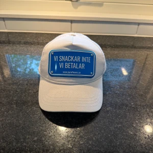 Vit keps från John Hatter & Co med blå skylt - Vit keps med mesh på sidorna och bak, framtill en blå skylt med texten 'VI SNACKAR INTE VI BETALAR'. Märke: John Hatter & Co. Justerbar passform och böjd skärm. Perfekt statement-accessoar.