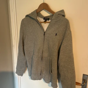 Grå hoodie från Polo Ralph Lauren - Grå ziphoodie med dragkedja från Polo Ralph Lauren. Klassisk design med huva, två fickor framtill och liten broderad logga på bröstet. Perfekt för en avslappnad stil.