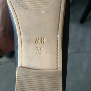 Svarta ballerinaskor med rosett från H&M - Säljer ett par svarta ballerinaskor från H&M i storlek 33. Skorna har en söt rosett framtill och ett elastiskt band över foten för extra komfort. Perfekta för dig som gillar en klassisk och stilren look.