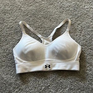 Vit sport-bh från Under Armour - Säljer en vit sport-bh från Under Armour med brottarrygg och elastiskt band under bysten. Bh:n har tunna axelband och är tillverkad i ett mjukt, stretchigt material som passar perfekt för träning.