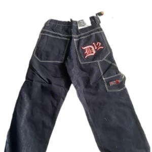 Svarta streetwear jeans med D12 brodyr - Säljer ett par svarta jeans från D12 streetwear med vit kontrastsöm och stor röd D12-brodyr på bakfickan. Byxorna har flera fickor och en loose passform, perfekta för en avslappnad stil. Väldigt små!